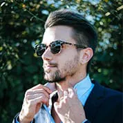 Man in a blazer and light blue shirt wearing sunglasses, adjusting his collar outdoors.